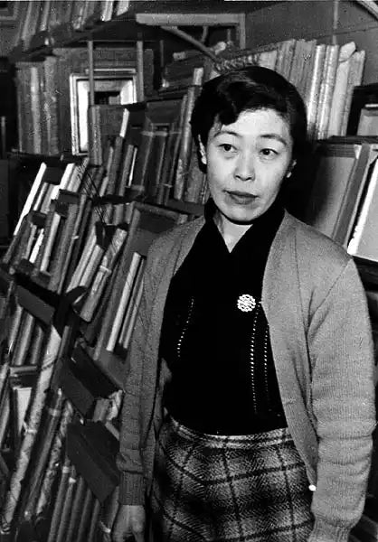 A woman with short, dark hair wearing a cardigan, a dark shirt with a brooch, and a plaid skirt.